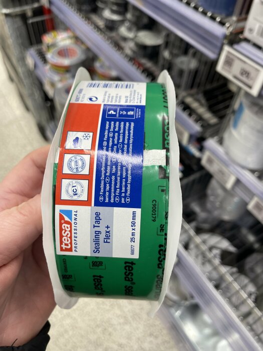 Rulle med grön byggtejp från Tesa, märkt "Sealing Tape Flex+", hålls i en hand i en butik.