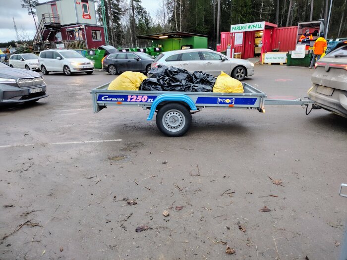 Släpvagn med svarta och gula sopsäckar på en återvinningsstation, omgiven av parkerade bilar och containrar för farligt avfall.