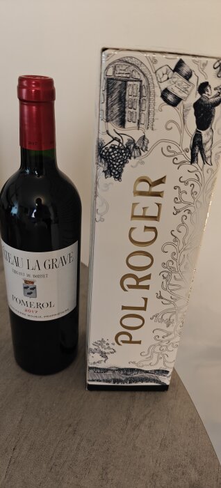 Vinflaska Chateau La Grave Pomerol 2017 och en förpackning Pol Roger champagne mot en ljus bakgrund på ett bord.