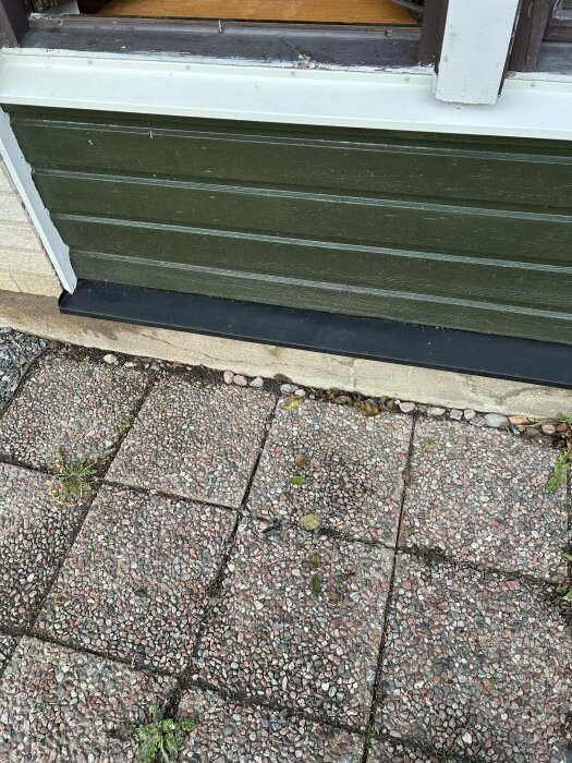 Fuktiga stenar framför en grön husvägg med tecken på fukt jämfört med torra plattor.