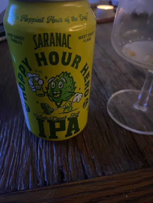 Burk med Saranac Hoppy Hour Hero IPA på ett bord, ett ölglas bredvid, mörk träyta och tänd ljuslykta i bakgrunden.