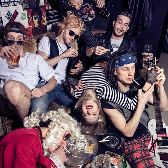 En grupp människor i rockstil sitter i en soffa med cigaretter och drinkar. En man med randig tröja spelar gitarr, och en person sover med huvudet i en peruk.