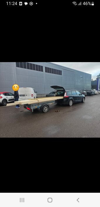 Bil med släpvagn lastad med brädor parkerad vid industribyggnad. Bakluckan är öppen och en person står bredvid.