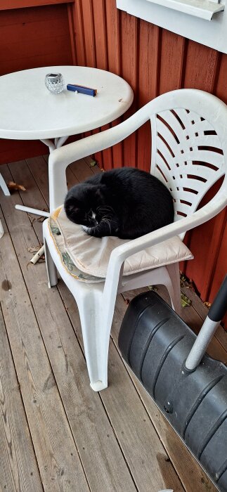 Svart katt ligger på en vit plaststol med kudde på en veranda, intill ett runt bord och en snöskyffel mot en röd vägg.