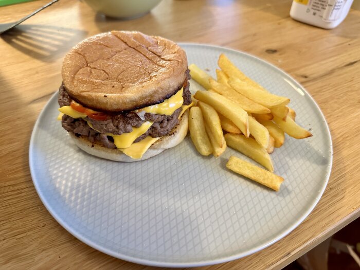 Hamburgare med smält ost och tomater serverad med pommes frites på en ljusgrå tallrik på ett träbord.