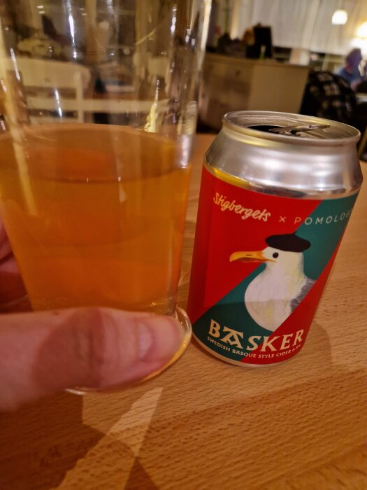 En burk Basker cider från Stigbergets med en illustration av en fågel, bredvid ett glas med cider, stående på ett träbord.