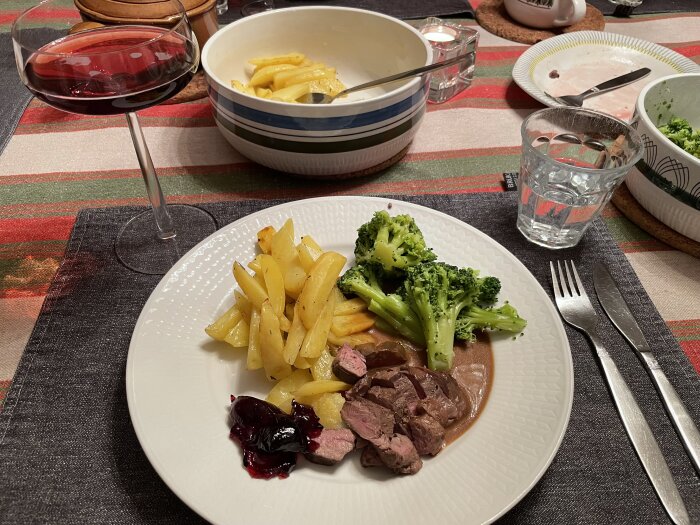 Tallrik med rådjursfilé, potatis och broccoli samt glas rödvin och skål med potatis på randig duk.