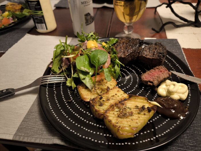 Tallrik med oxfilé, accordion potatoes, sallad och två olika såser. Bakgrund med ölfat och glas.