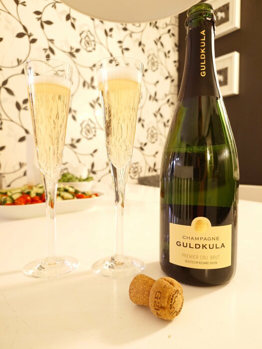 Två glas med svenskt mousserande vin Guldkula och en flaska på ett bord, med en kork och ljus bakgrund.