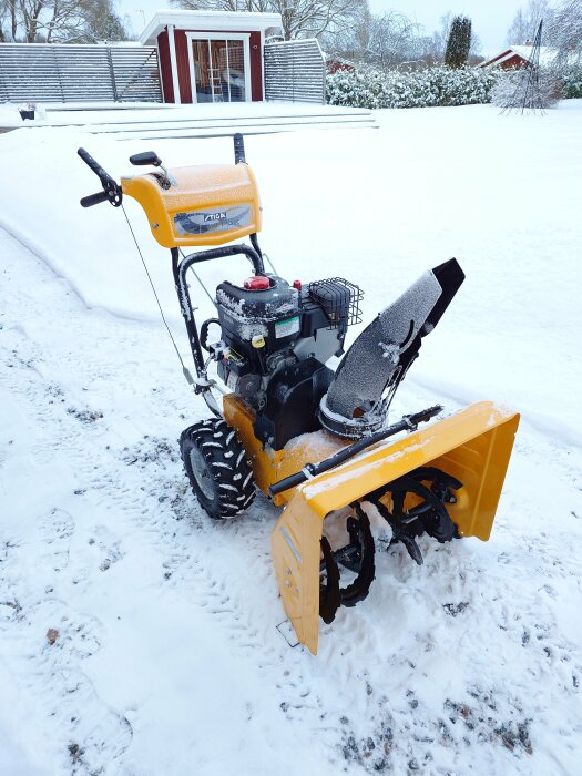 Gul snöslunga i en snötäckt trädgård framför en röd byggnad, används för att hantera 1 dm pudersnö. Saknar belysning.