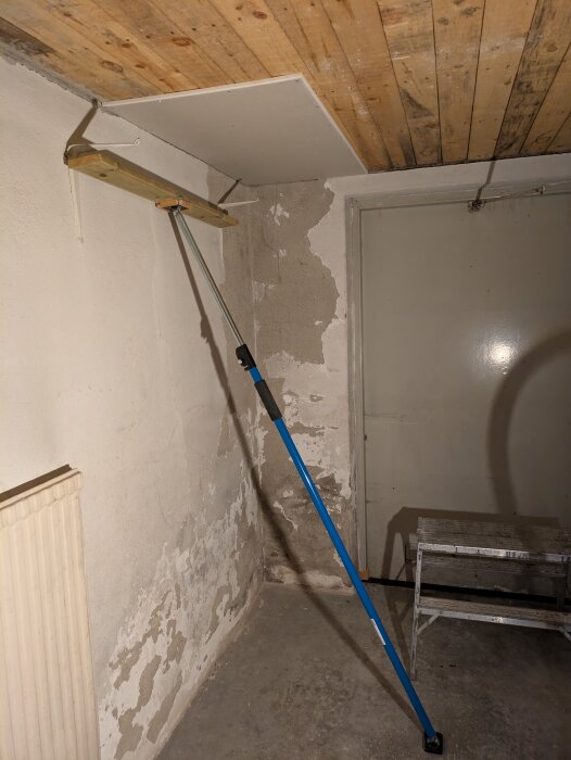 Gipsskiva uppsatt i taket med hjälp av stöttstativ och trästöd i ett rum under renovering, avskalade väggar och en dörr i bakgrunden.