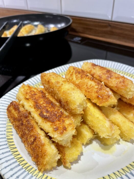 Panerad halloumi på en tallrik, gyllenbruna och krispiga, med ytterligare bitar som steks i en panna i bakgrunden.