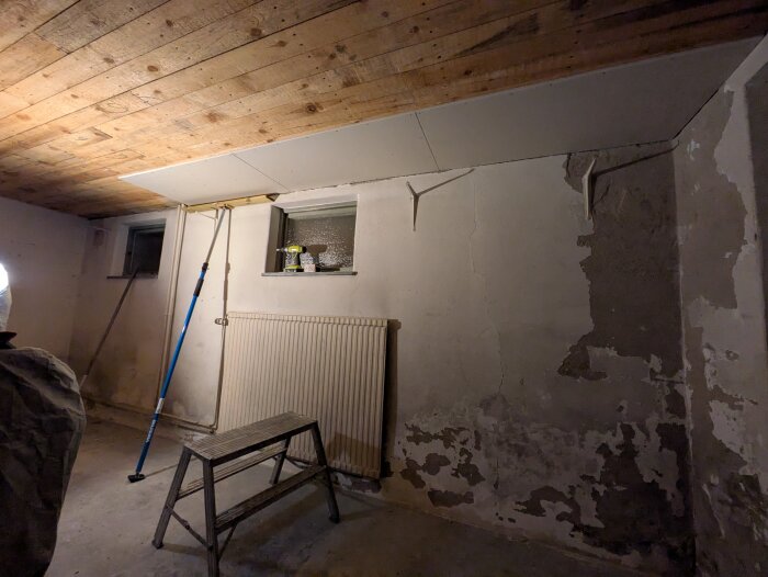 Rum med påbörjad montering av takgips, ett hopfällbart arbetsbord och en fuktig vägg som behöver dräneras innan putsning.