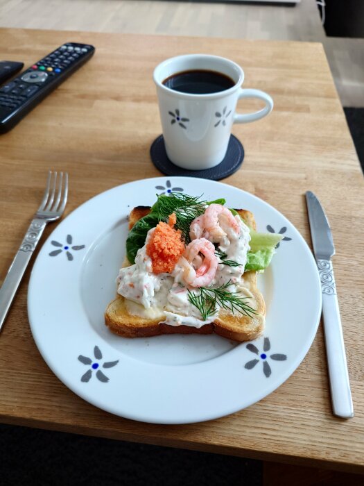 Skagen toast med dill, räkor och caviar på en tallrik, bredvid en kopp kaffe och bestick på ett träbord.