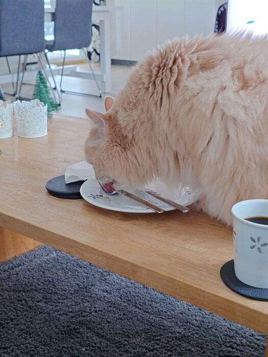 En ljus katt slickar på en tallrik på ett träbord, bredvid ligger bestick och en kopp kaffe.