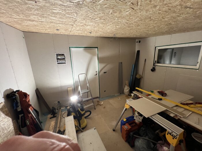 Rum under renovering med uppsatta gipsskivor, stege, verktyg och byggmaterial på golvet och arbetsbordet.