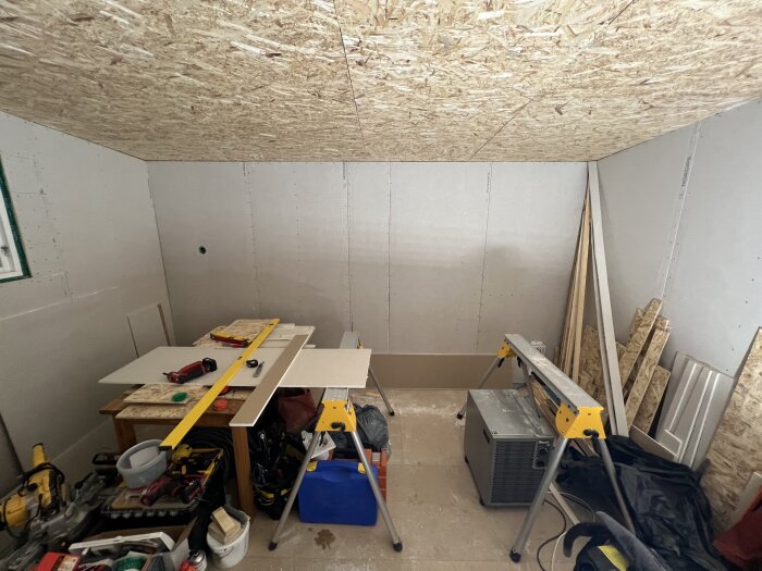 Ett rum med nygipsade väggar och osb-skivor i taket, verktyg och byggmaterial syns på golvet.