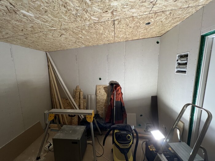 Rum med väggar klädda i gipsskivor, OSB-skiva i taket, byggmaterial och verktyg på golvet, inklusive en dammsugare och stege.