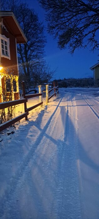 Snöig uppfart med hjulspår vid rött hus i kvällsljus, trästaket och bar trädgren. Mörkblå himmel och belysning från huset.