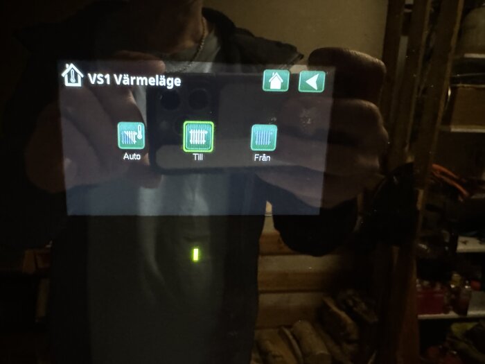 Skärm som visar värmeläge med alternativen "Auto", "Till" och "Från" aktiverade på ett värmesystem. Reflektion av person syns.