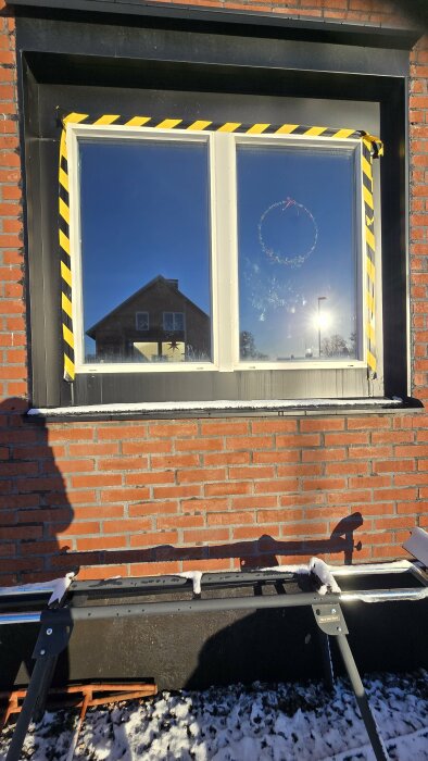 Stort fönster med gulsvart tejp runtom, mot röd tegelvägg. Fönstren reflekterar ett hus och solen. Tejp förhindrar vattenläcka.