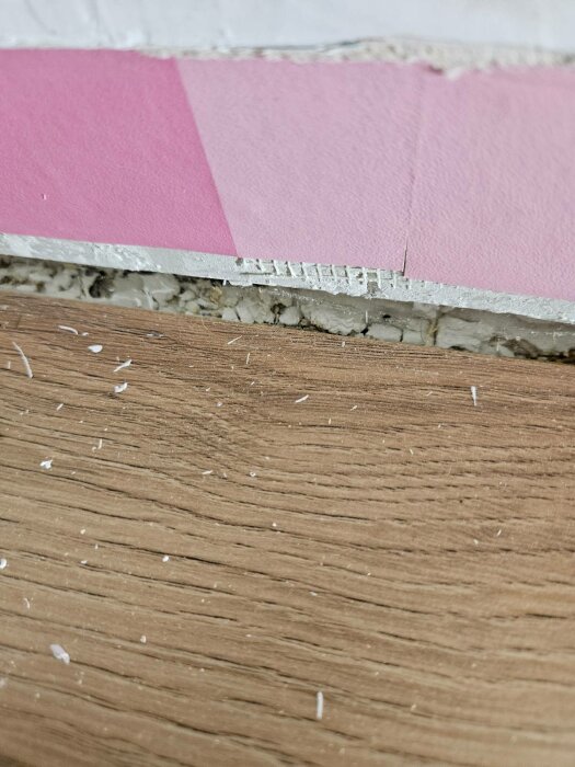 Närbild av rosa tapet och trälist med exponerat vitt poröst material i väggen, potentiellt misstänkt för asbest eller gips.