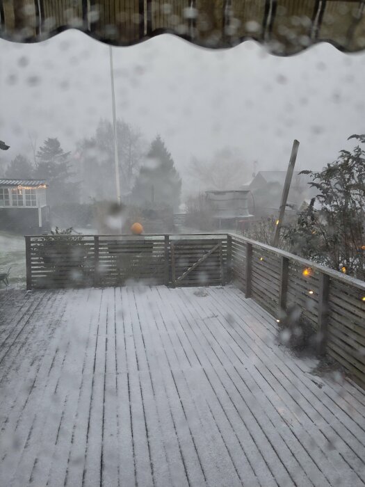 Utsikt från ett fönster över en altan som är täckt av ett lager snö, dimmig bakgrund med träd och byggnader.