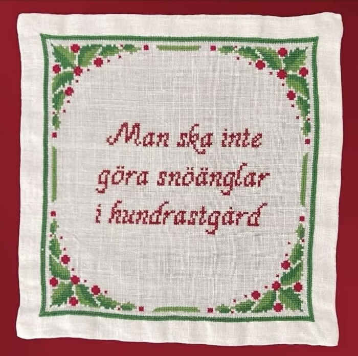 Broderad duk med texten "Man ska inte göra snöänglar i hundrastgård" omgiven av gröna och röda dekorativa mönster på en röd bakgrund.