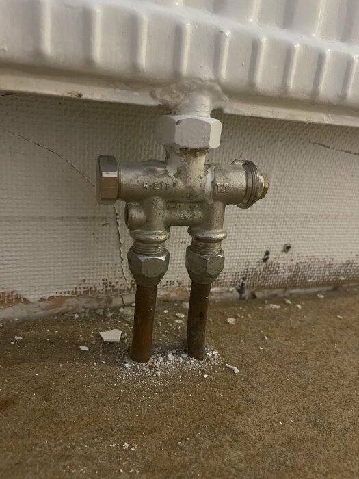 Närbild av radiatoranslutningar med ventiler och insexnyckel, installerade på en vägg i en källare. Finns byggdamm på golvet under.