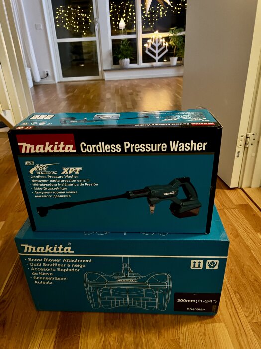 Två Makita-produkter; en sladdlös högtryckstvätt och ett snöslungsfäste i sina originalförpackningar på ett trägolv.