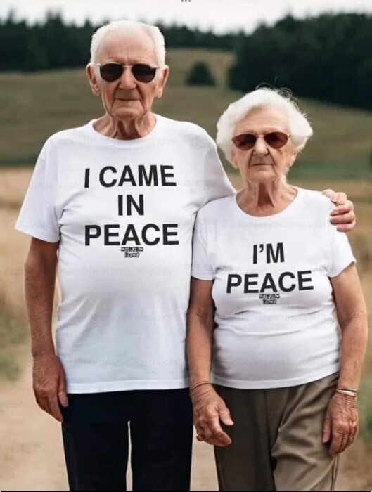 Äldre par i vita t-shirts med texten "I CAME IN PEACE" och "I'M PEACE", står tillsammans på en landsväg med skog i bakgrunden.