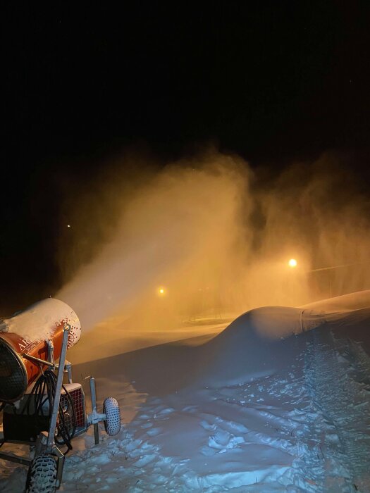 Snökanon sprutar snö i natten, med upplysta snöhögar och fallande snöflingor i bakgrunden.