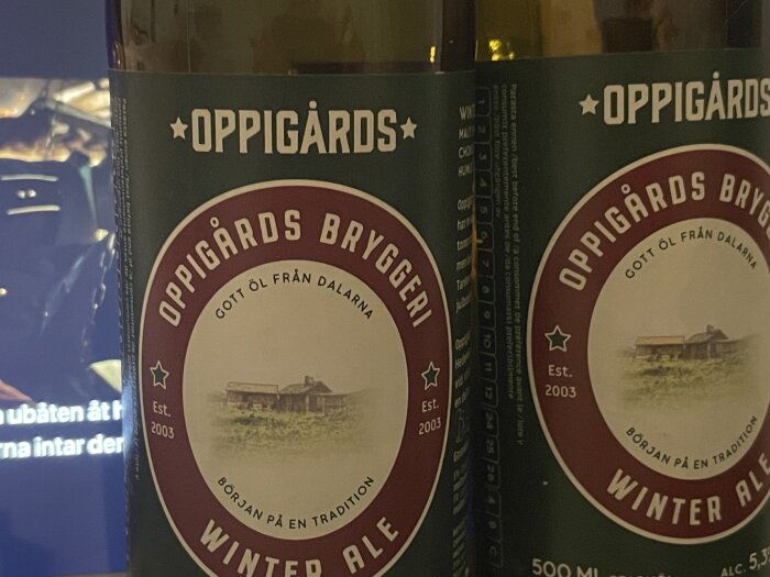 Flaskor med Oppigårds Winter Ale, bryggda i Dalarna. Flaskorna står framför en skärm, etiketter i fokus med en bondgård i bild.