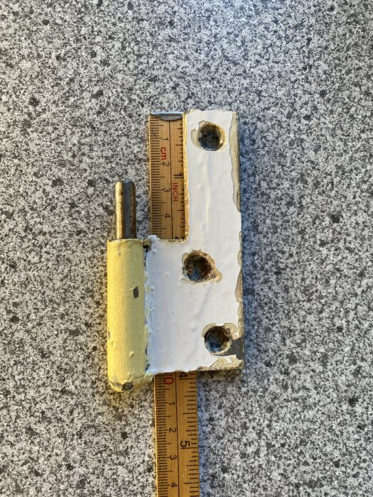 Gångjärn med vit färg, placerat bredvid en linjal på grå yta, visar skala i centimeter och tum.