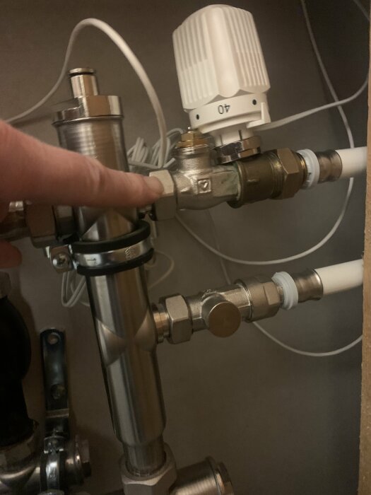 Finger pekar på ett rör vid en koppling i ett VVS-system, termostat sitter intill och kablar syns i bakgrunden.