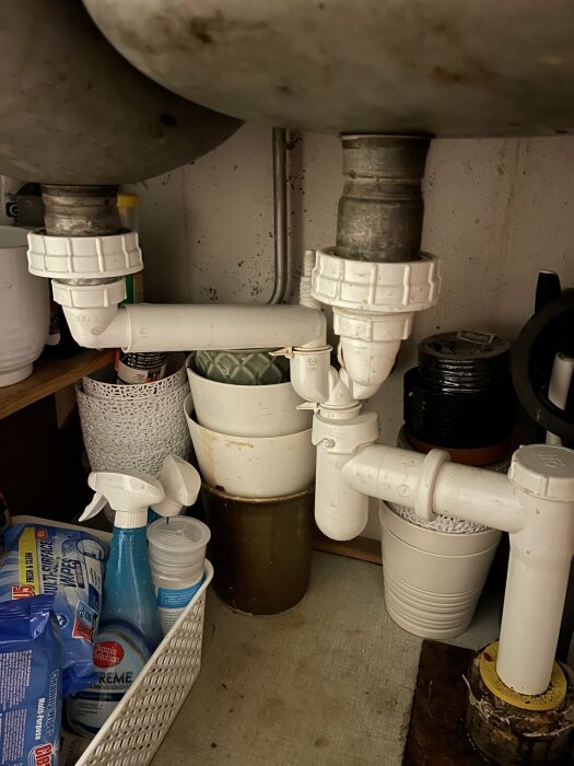 Ett gammalt vattenlås under diskbänk med läckande kopplingar, omgiven av städprodukter och krukor i ett köksskåp.