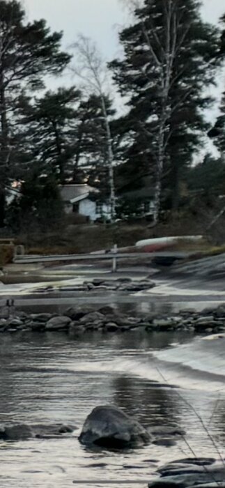 Klippa vid sjön kantad av träd, med stilla vatten och stenblock i förgrunden.