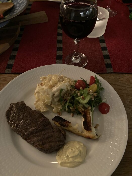 Tallrik med kött, sallad, potatisgratäng, bröd och sås samt ett glas rödvin.