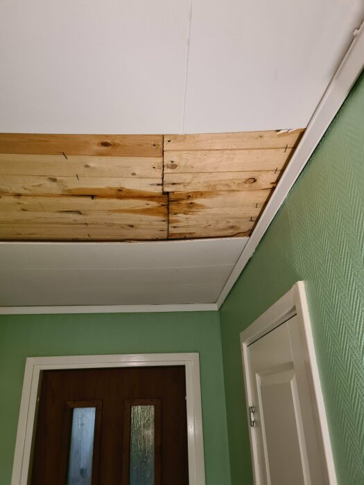 Skadat innertak med borttagna paneler som visar fuktfläckar och trä i en grönmålad hall.