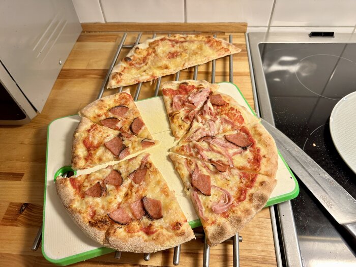 Hembakad pizza på skärbräda, toppad med skinka och korv, ligger i bitar på köksbänk. Pizzan bakades utan fungerande ugn.