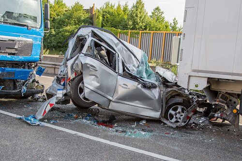 Sönderkrossad silverfärgad bil klämd mellan två lastbilar på motorväg, omgiven av krossat glas och skräp, med skog i bakgrunden.