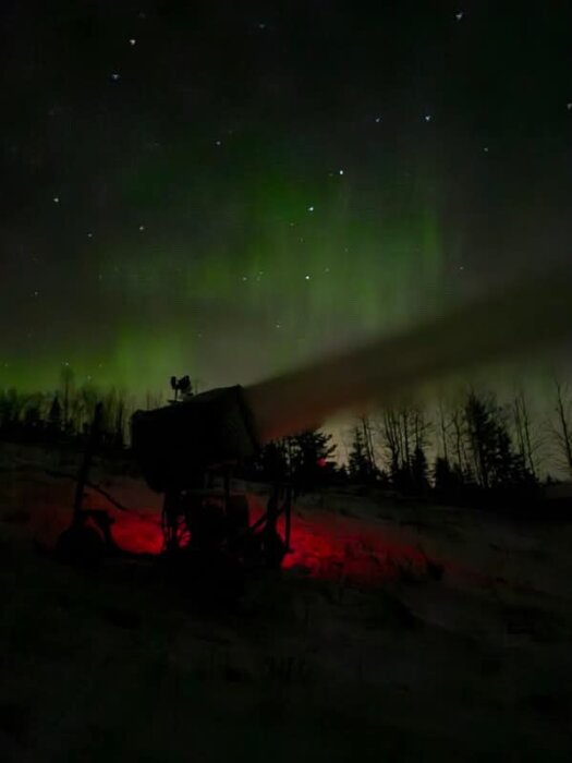 Snökanon under natthimmel med grönt norrsken och stjärnor, belyst av rött ljus vid basen, omgiven av träd i bakgrunden.