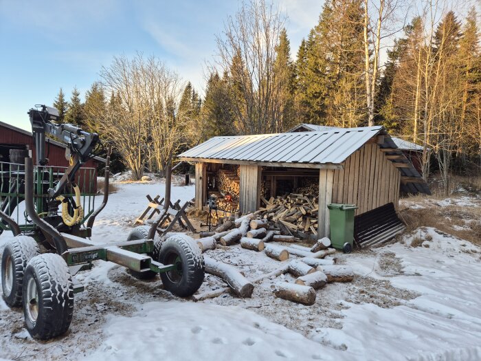 Vedtrave utanför en träbod i vintermiljö med snö på marken. Ett släpvagnsstativ finns vid sidan av boden.