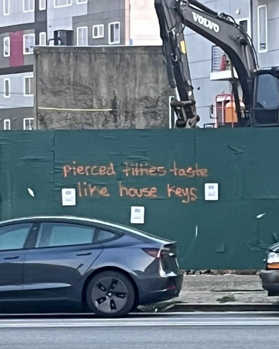 Grön byggnadsstängsel med orange graffiti som säger "pierced titties taste like house keys", i bakgrunden syns en Volvo-grävmaskin och bostadshus.