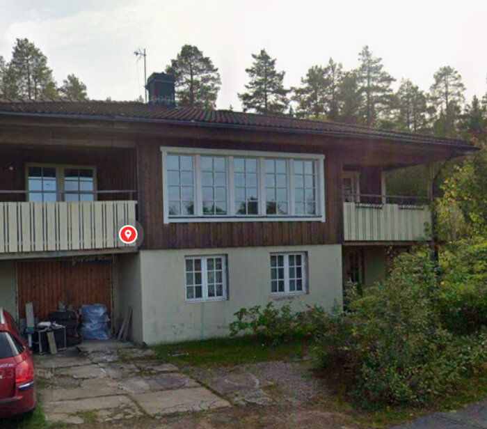 Sutterängvilla från 1980-talet med putsad nedre del och laserat trä på övre våningen, fotad från Google Maps, omgiven av träd och med en balkong.