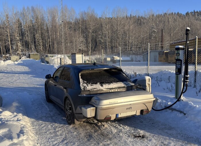 Hyundai Ioniq 5 laddas vid en laddstation i snötäckt vinterlandskap, med snö på bilen och omgivningen under solig himmel.