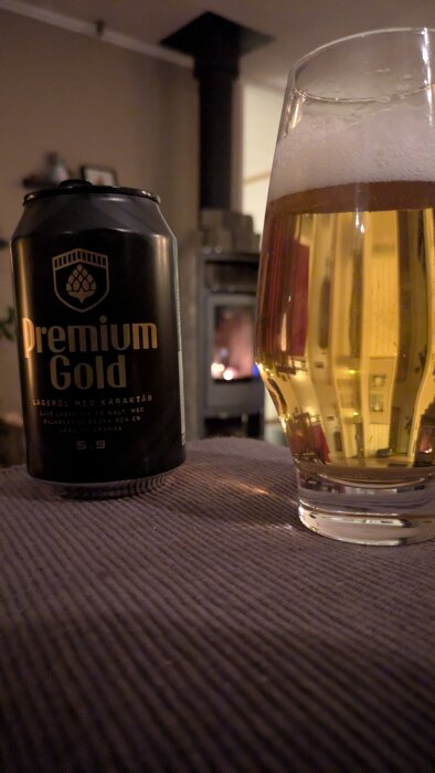 Burk och glas med ljus lageröl Premium Gold, placerade bredvid en tänd kamin i ett mysigt hemönskemiljö.