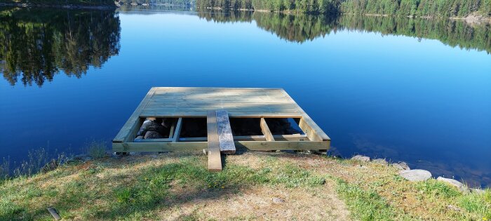 Nybyggd brygga av trä vid en sjö, reflektioner av träd i vattnet.