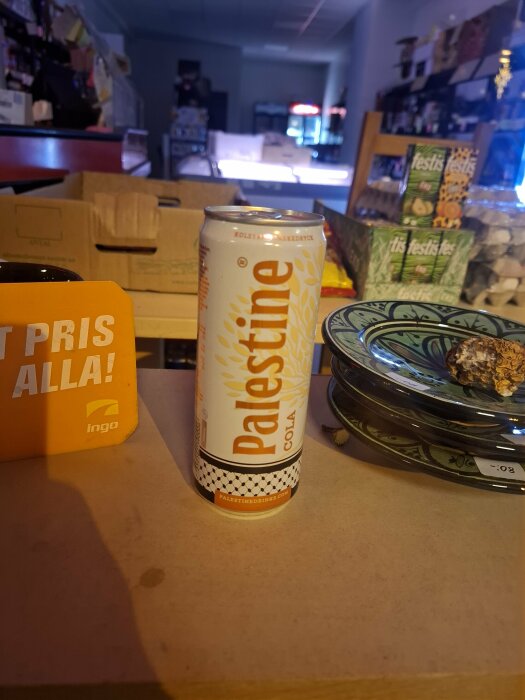 Burk med Palestine Cola står på ett bord i en butiksmiljö, bredvid tallrikar och skylt med texten "Toppris för alla!".