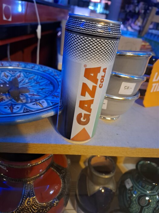 Burk med Gaza Cola placerad på hylla bredvid färgglada keramikplattor och metallburkar i en butik.